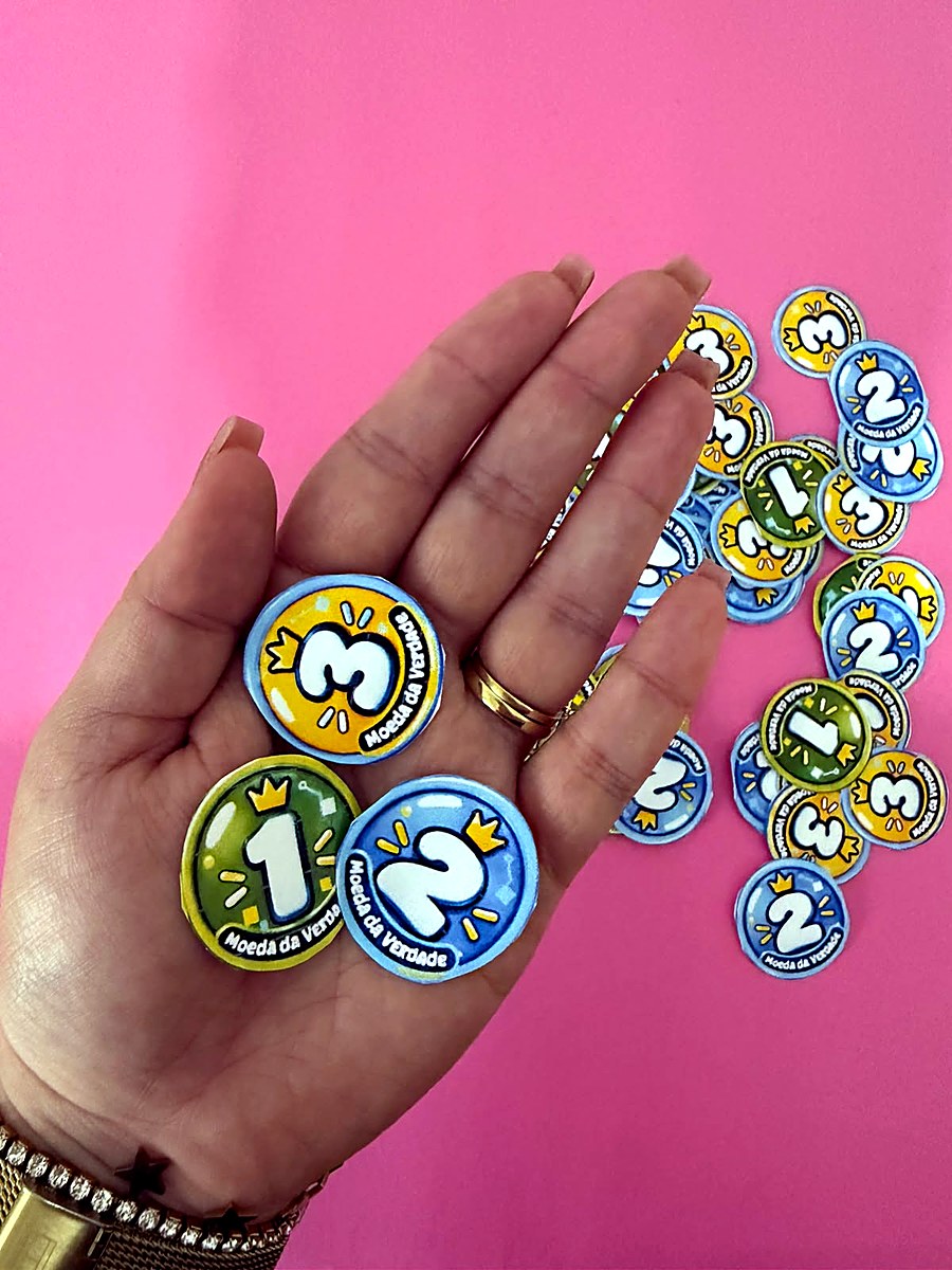 Foto real de uma mão segurando as Moedas da Verdade de 1, 2 e 3 pontos, com outras moedas sobre a mesa